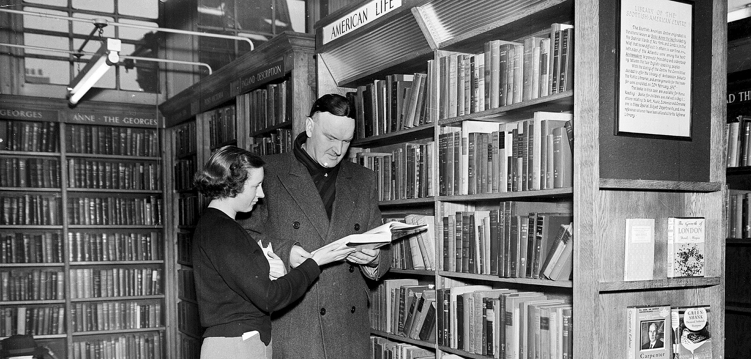 A black and white photo of a woman and man in a library looking at a book