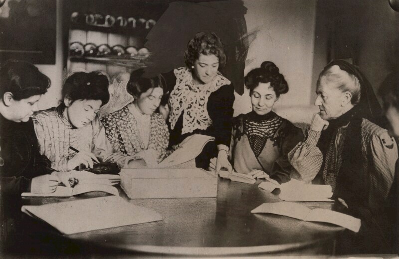 A group of women gathered around a table reading papers. Image © National Portrait Gallery, London. Attribution-NonCommercial-NoDerivs 3.0 Unported (CC BY-NC-ND 3.0)